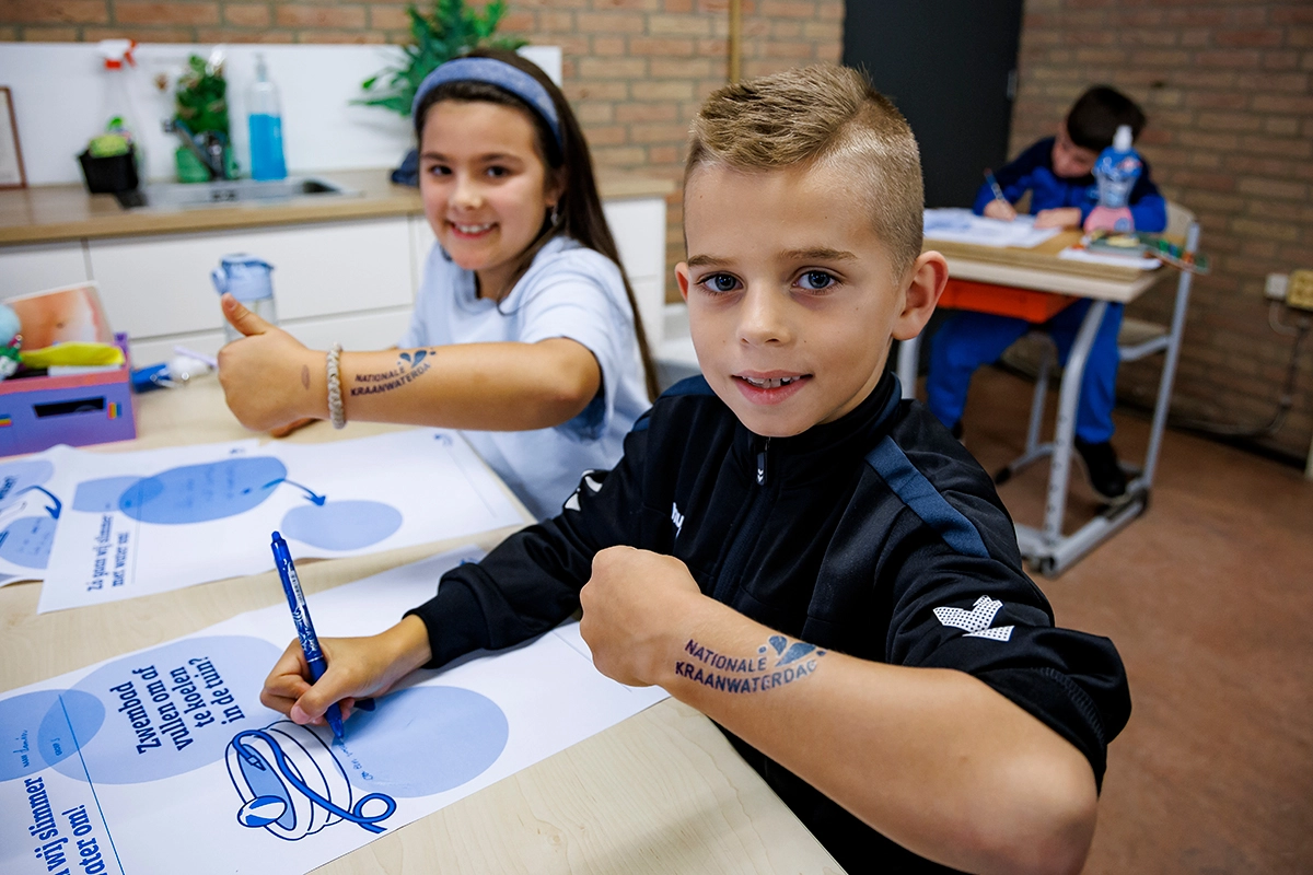Twee kinderen tonen tijdelijke plaktattoos met “Nationale Kraanwaterdag” op hun arm terwijl ze werkbladen invullen.