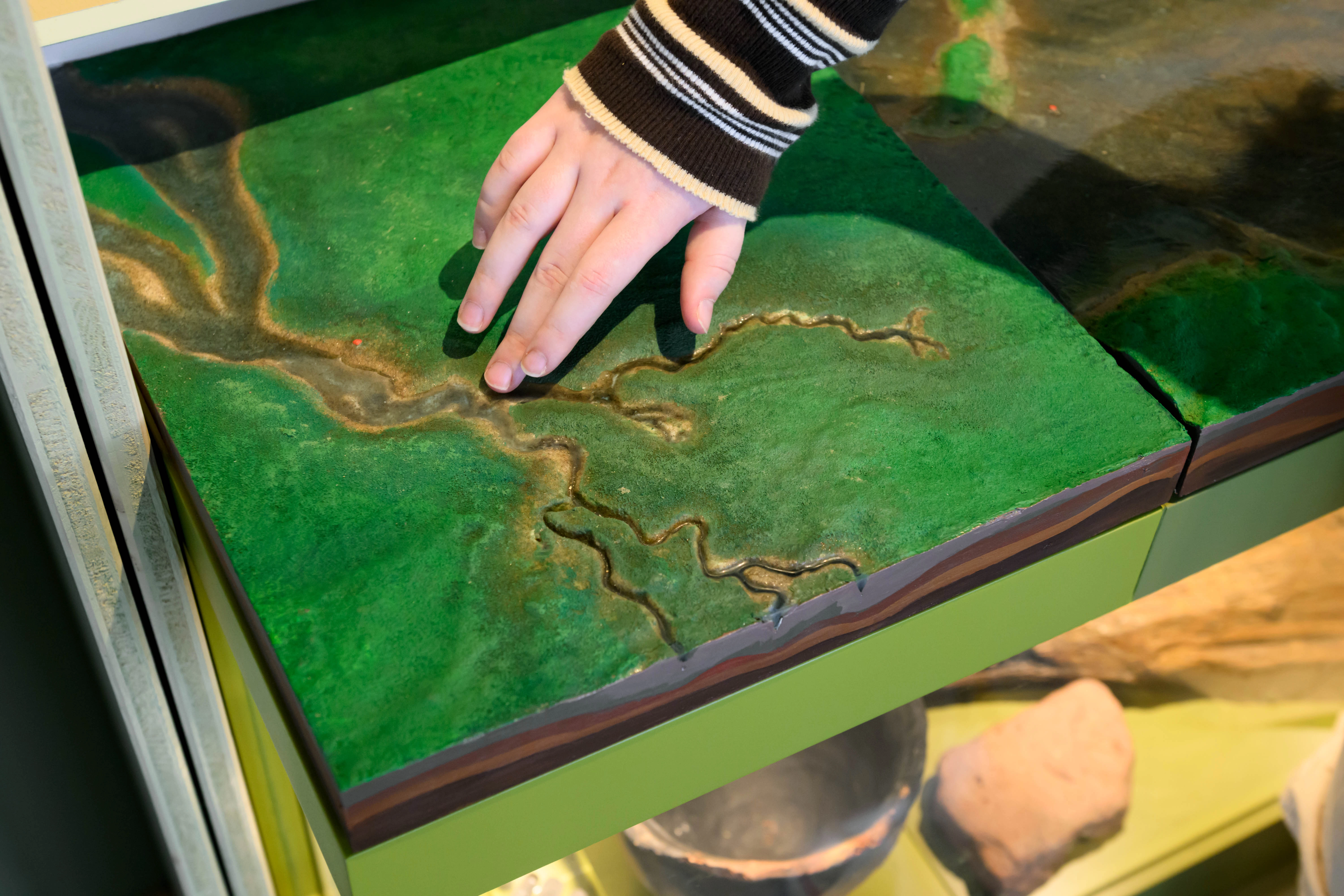 Close-up van handen die maquette met rivieren en valleien verkennen, onderdeel van leerzame expo over natuurlijke processen.