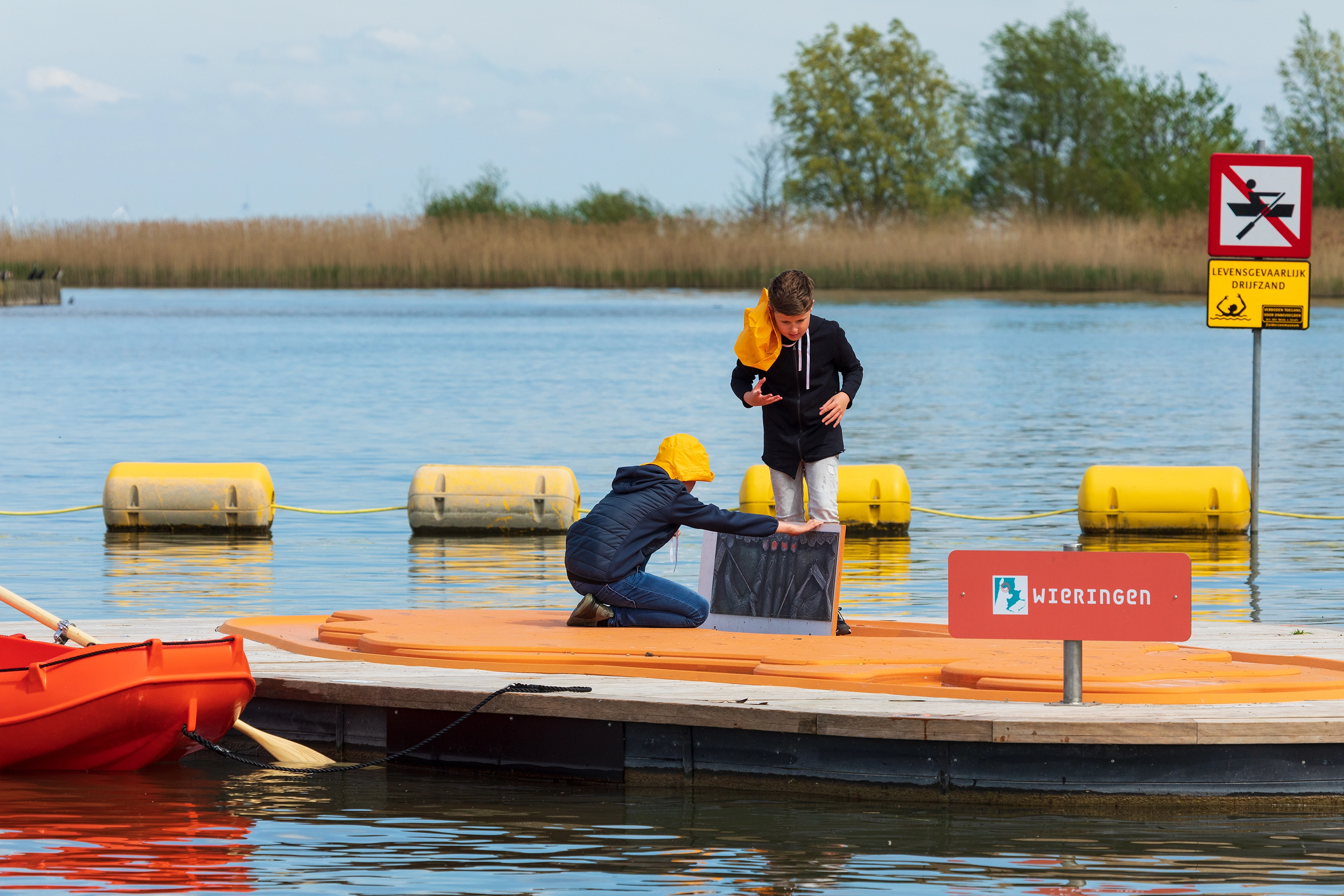 Twee kinderen op een vlot met het bord “Wieringen”, inspecteren een zwart bord terwijl hun roeiboot stil ligt.