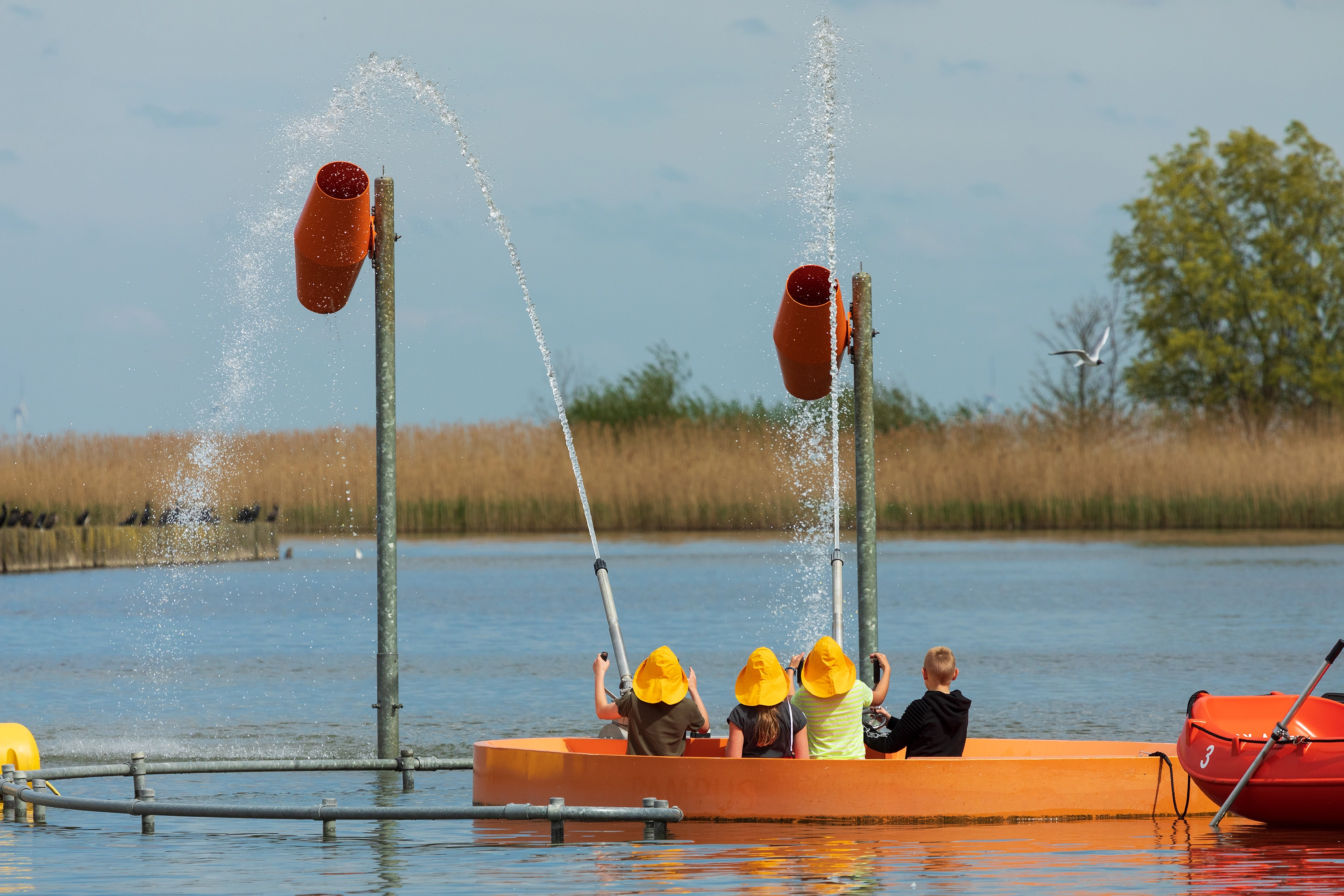 Drie kinderen spuiten water uit kanonnen vanaf een oranje platform in het meer, vlakbij een rietkraag.