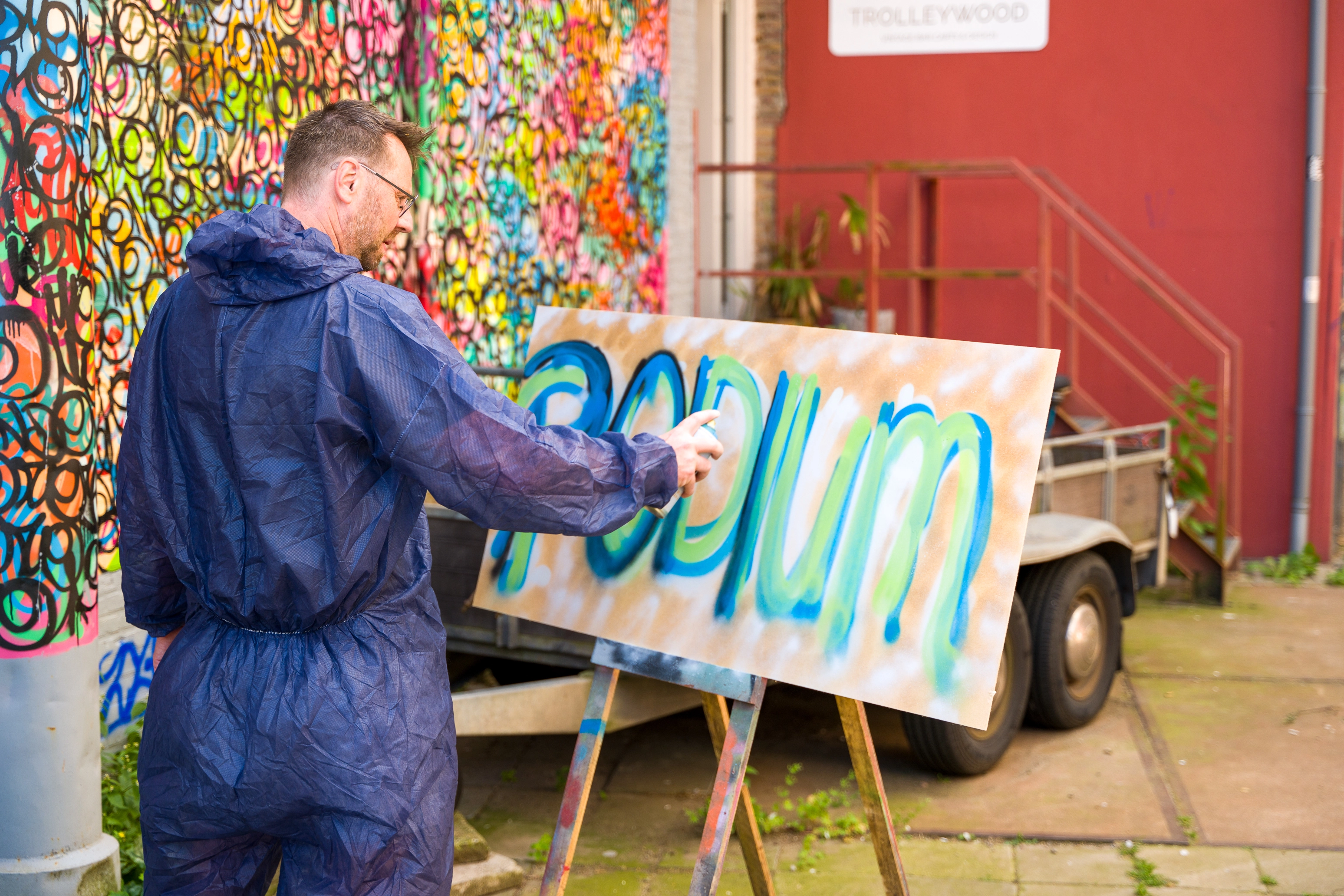 Persoon in overall spuit het woord “Podium” in blauwe en groene tinten op een paneel, kleurrijke muur achter hem.