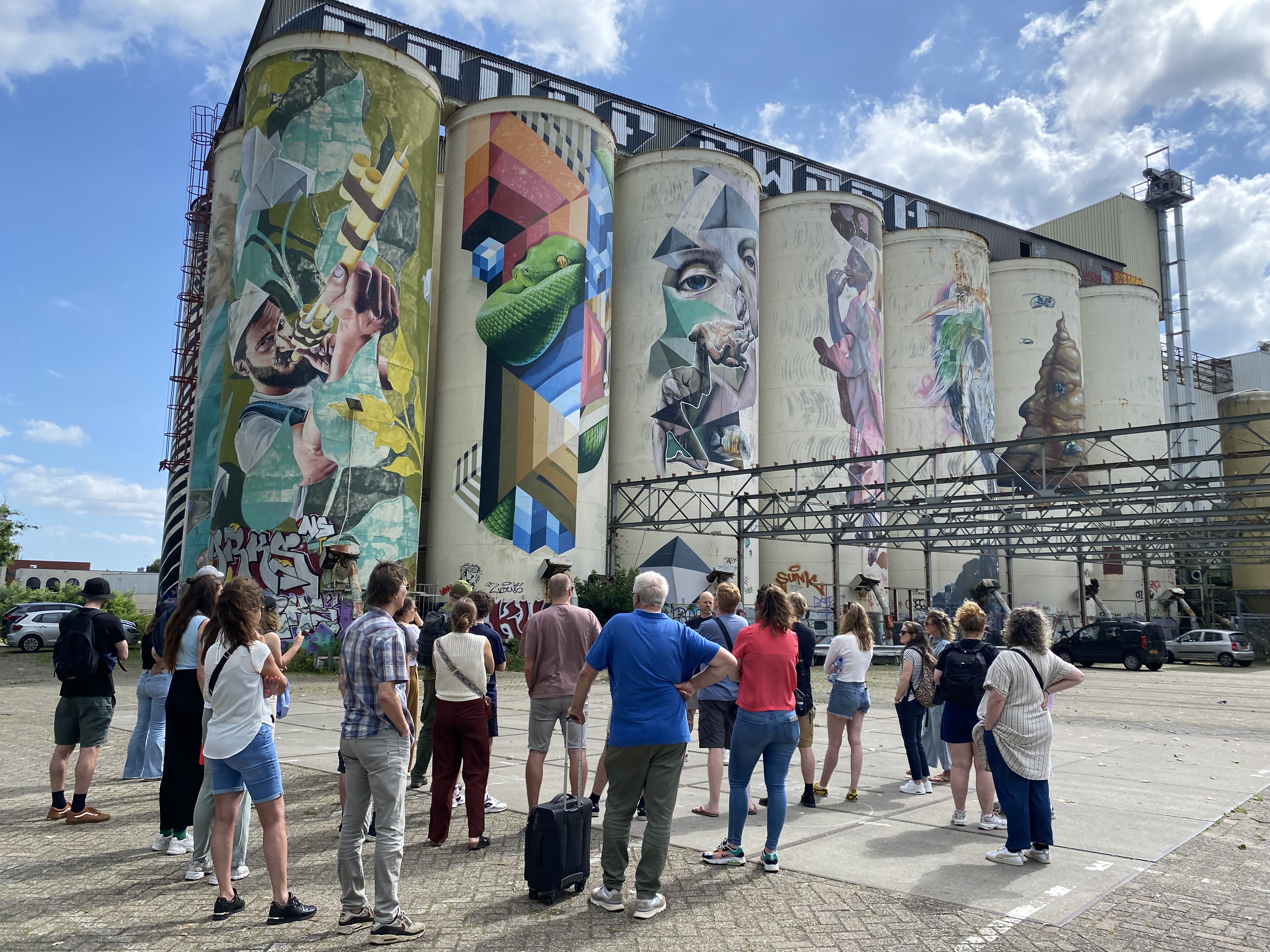 Groep bekijkt grote silo’s met indrukwekkende muurschilderingen van dieren en abstracte vormen op industrieterrein.