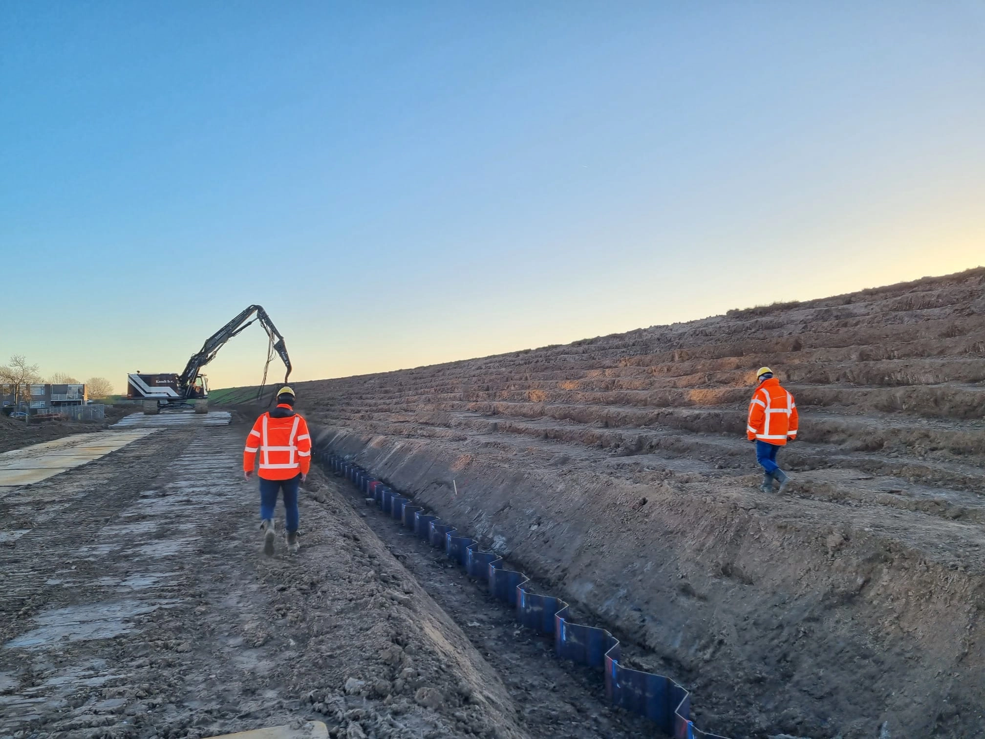 Twee medewerkers lopend op een dijk. Achterin het beeld is een graafmachine bezig met dijkversterking.