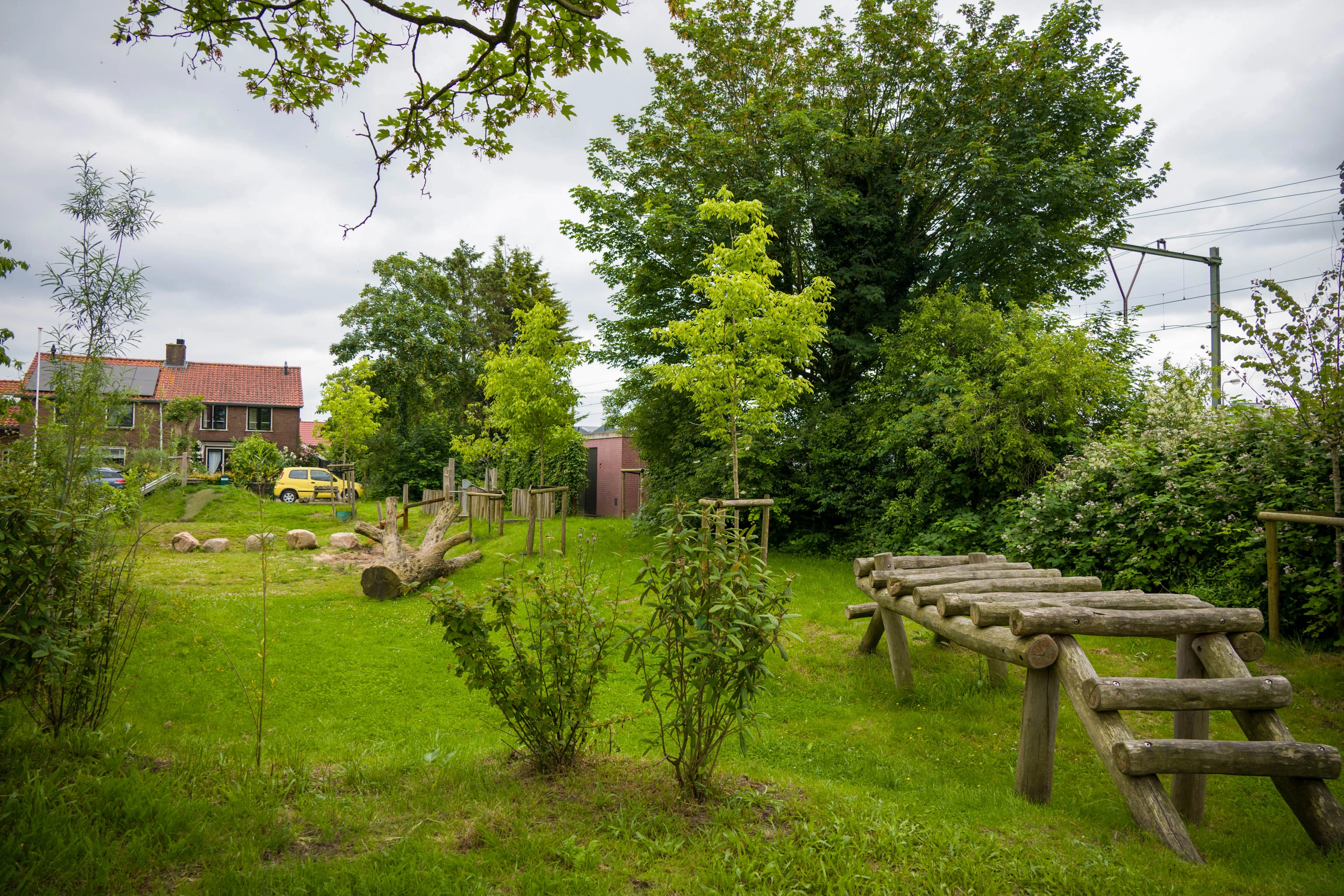 Een groene speelplaats in een woonwijk met veel gras, bomen en houten speeltoestellen