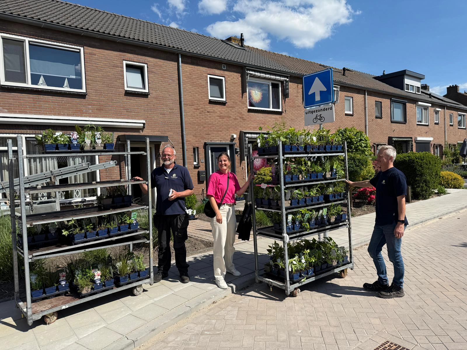 Er worden plantjes in de straat uitgedeeld door de omgevingsmanager en de gemeente. Je ziet twee mannen en een vrouw in bij een kraam met plantjes staan.